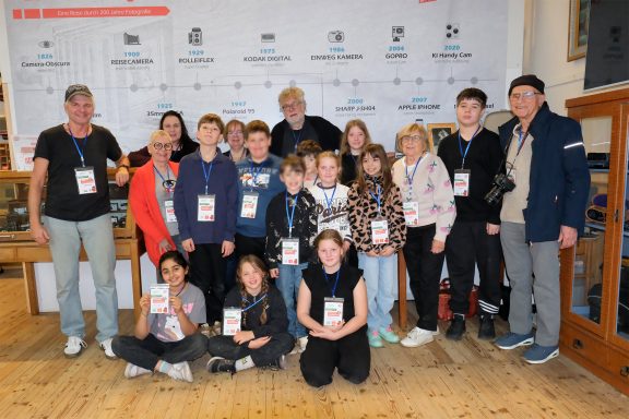 Gruppenfoto von Kindern und Erwachsenen mit Ausweisen, lächelnd vor einer Wand.