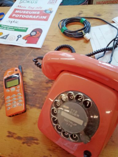 Roter Vintage-Telefon und orangefarbener Handy auf Holztisch, daneben Zeitschrift.