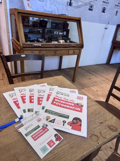 Informationsmaterial und ein Schreibwerkzeug liegen auf einem Tisch vor einer Vitrine.